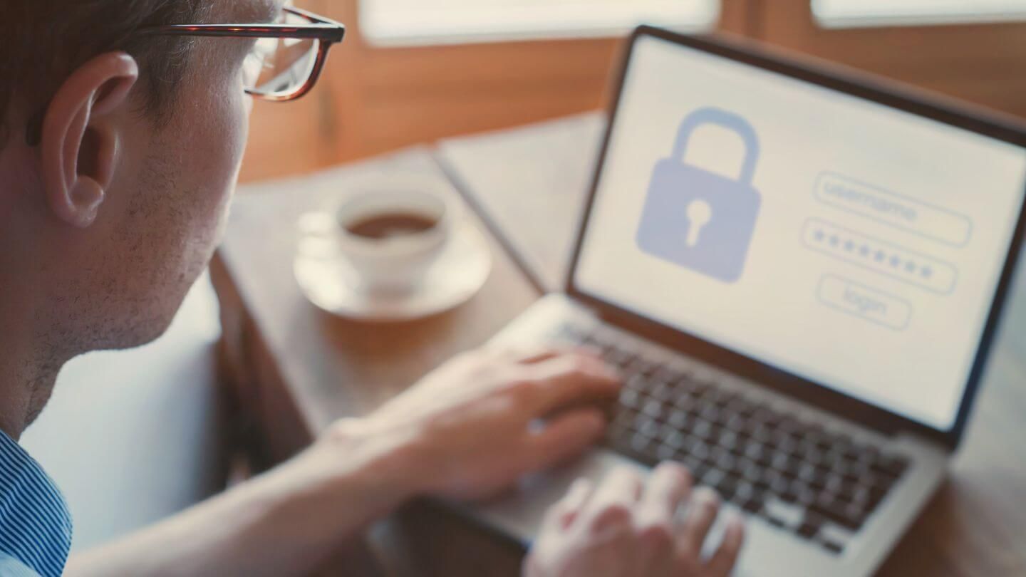 Man with glasses sitting at a laptop displaying a login screen with a lock icon and password fields.