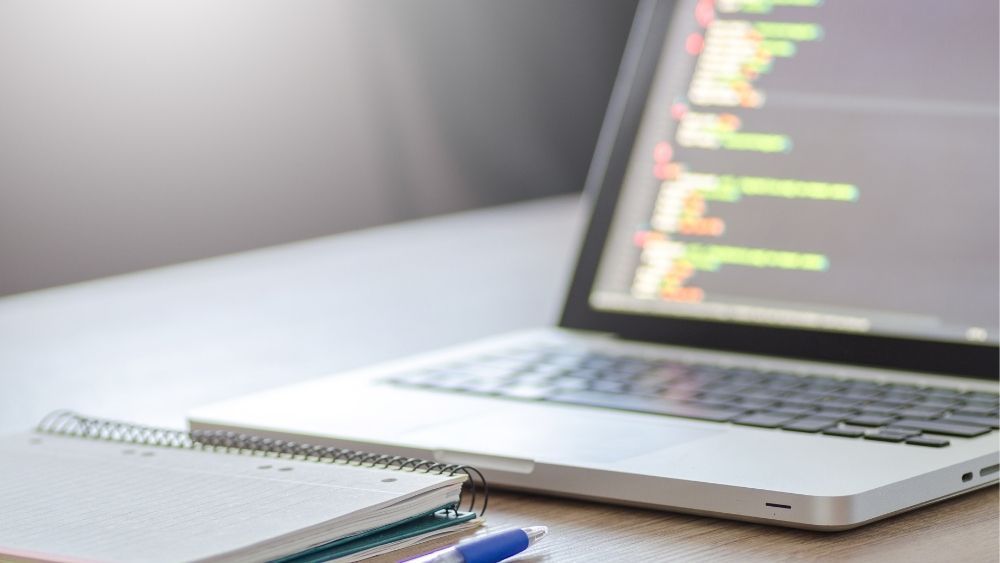 Laptop with lines of code sitting on a desk with a notepad and pen